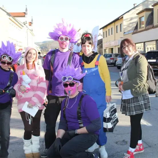 Fünf Personen in bunten Kostümen und Gesichtsbemalung stehen auf einer Straße. Eine hält einen weißen Plüschtier, und eine andere trägt eine Brille. Hinter ihnen sind Gebäude und geparkte Autos.