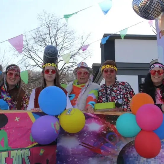 Eine Gruppe von Menschen in 70er-Jahre-Kleidung steht auf einem Wagen mit Luftballons. Sie lächeln und tragen Sonnenbrillen. Der Wagen hat einen Banner, auf dem 'Die 70er' steht.