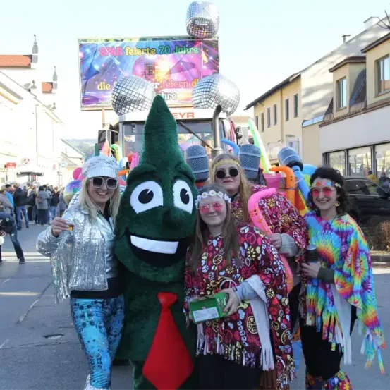 Fünf Personen posieren für ein Foto vor einer Discokugel auf einem Wagen. Eine hält einen grünen Maskottchen. Dahinter versammelt sich eine Menschenmenge, und Gebäude säumen die Straße.
