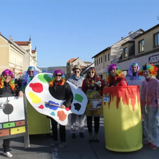 Eine Gruppe von Menschen in bunten Kostümen, einschließlich Perücken und Pinseln, posiert für ein Foto auf einer Straße, gesäumt von Gebäuden und einem klaren Himmel.