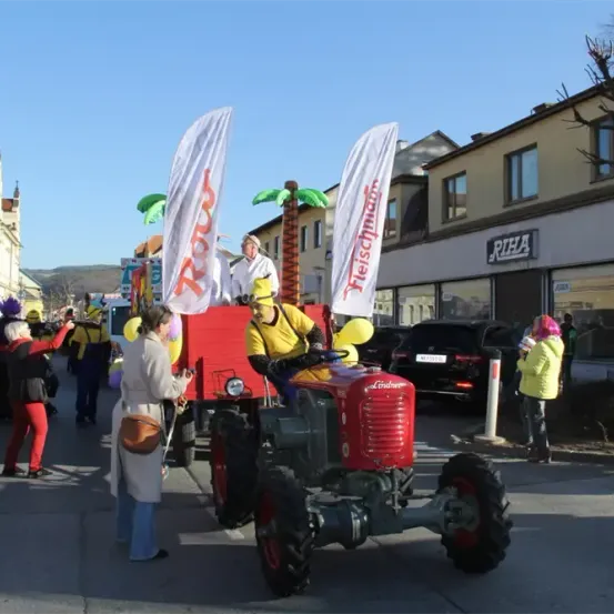 Ein roter Traktor mit Bannern fährt mit Leuten in Kostümen, Luftballons und einer Frau in einem weißen Mantel durch die Straße. Dahinter befindet sich ein Gebäude mit einem Schild, auf dem RIHA steht.