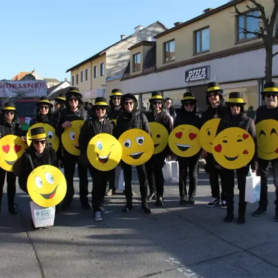 Eine Gruppe von Menschen in Schwarz gekleidet, mit Sonnenbrillen und Hüten, posiert für ein Foto und hält große Ausschnitte von Smiley-Gesichtern. Sie stehen vor einem Gebäude mit einem Schild, auf dem 'RIFA' steht.
