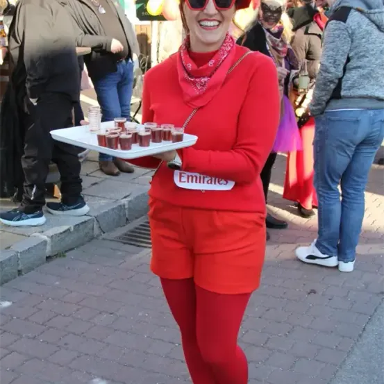 Eine Frau in rotem Outfit trägt einen Tablett mit Getränken bei einem Karneval, trägt eine Sonnenbrille und einen roten Hut. Hinter ihr sind Menschen in verschiedenen Kostümen gekleidet, darunter eine Person mit einer Maske.