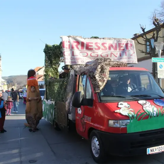 Ein roter Lastwagen mit einem dekorierten Frontteil, das Tieraufkleber und ein Banner mit der Aufschrift 'Griessner Gloggnits' zeigt, fährt während eines Festivals eine Straße entlang. Menschen beobachten, und ein Mann in einem braunen Kostüm steht neben dem Lastwagen.