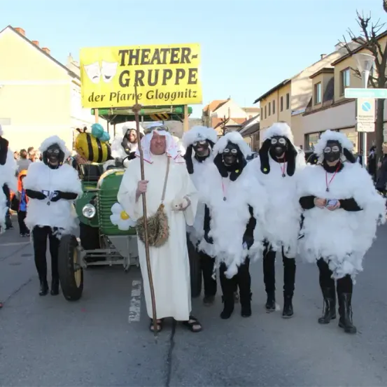 Eine Gruppe von Menschen in weißen Kostümen mit schwarzen Gesichtern nimmt an einem Umzug teil. Ein Mann in einer weißen Robe steht vor ihnen. Hinter der Gruppe befindet sich ein Fahrzeug mit einem gelben Schild, auf dem 'Theater-Gruppe der Pfarre Gloggnitz' steht. Gebäude befinden sich am Straßenrand.