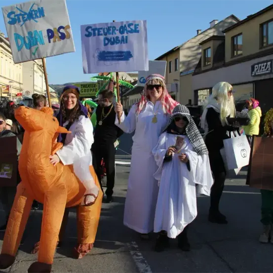 Eine Gruppe von Menschen in Kostümen läuft bei einer Parade. Eine Person reitet auf einem orangefarbenen aufblasbaren Kamel. Sie halten Schilder mit der Aufschrift Steuer-Oase Dubai.