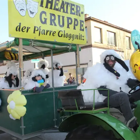 Eine Theatergruppe, 'der Pfarre Glognitz,' zieht in einem grünen Traktor mit einem gelben Schild, mit Schafsmaskottchen und einer Person in einem Schafskostüm mit einem gelben Ballon.