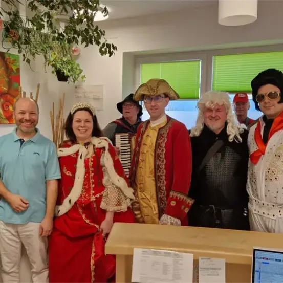 Eine Gruppe von Menschen in verschiedenen Kostümen posiert für ein Foto in einem Büro. Der Mann in Blau steht neben einer Frau in Rot. Hinter ihnen spielt ein weiterer Mann Akkordeon.