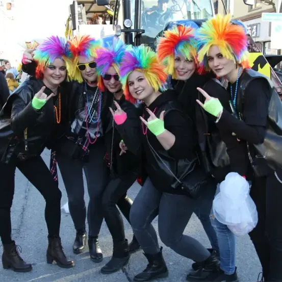 Sechs Frauen mit bunten Perücken posieren für ein Foto vor einem Fahrzeug. Sie tragen schwarze Jacken und Hosen. Sie alle lächeln und machen Friedenszeichen.