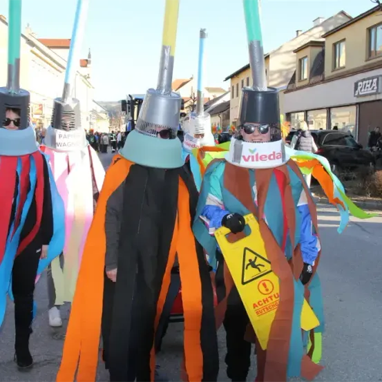 Eine Gruppe von Menschen in farbenfrohen Kostümen mit großen silbernen Hüten und Sonnenbrillen läuft auf der Straße. Einer von ihnen hält ein gelbes Schild mit einem Warnsymbol.