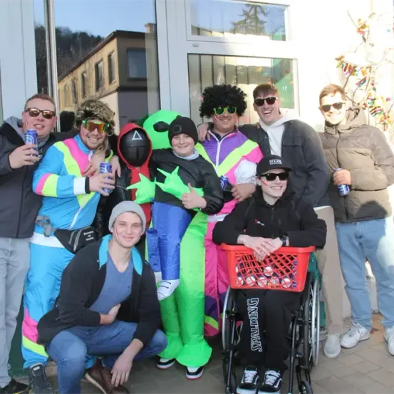 Eine Gruppe von Menschen in farbenfrohen Kostümen posiert für ein Foto. Eine Person sitzt im Rollstuhl mit einem Korb voller Dosen. Sie stehen vor einem Gebäude mit Glasfenstern.