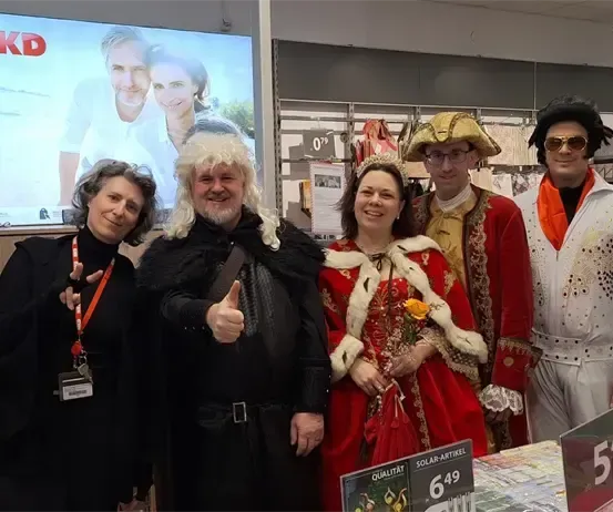 Eine Gruppe von Menschen in verschiedenen Kostümen posiert für ein Foto. Sie stehen vor einem Tisch mit zu verkaufenden Artikeln. Dahinter zeigt eine Projektionsleinwand ein Bild von einem lächelnden Mann und einer Frau.