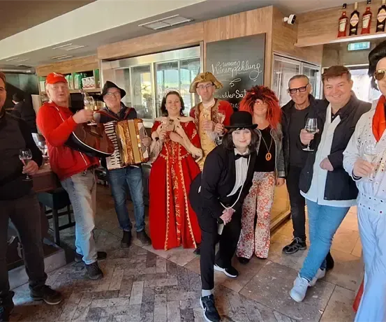 Eine Gruppe von Menschen in Kostümen posiert in einer Bar, einige halten Weingläser und ein Akkordeon.