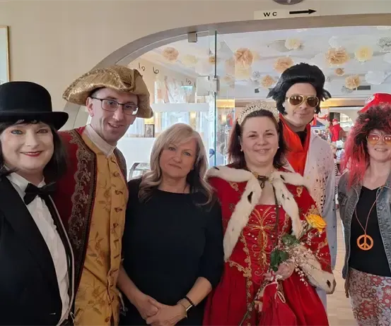 Eine Gruppe von Menschen in Kostümen posiert für ein Foto in einem dekorierten Raum mit einem Hinweisschild für die Toilette im Hintergrund.