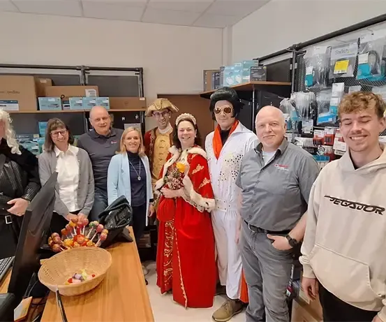 Mehrere Personen posieren für ein Foto in einem Raum mit einem Schreibtisch, einem Monitor und einem Korb mit Süßigkeiten. Einige tragen Kostüme.