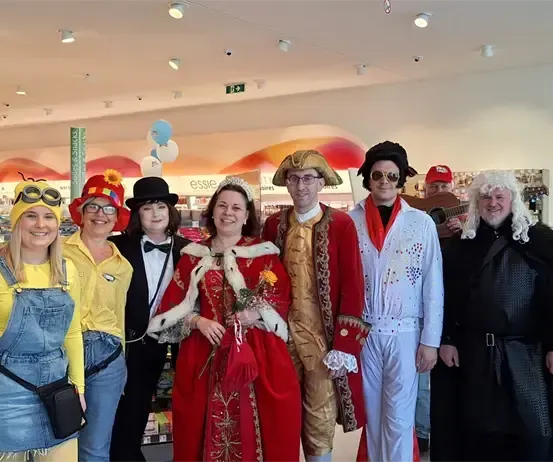 Eine Gruppe von Menschen in verschiedenen Kostümen steht in einem Geschäft, lächelt und posiert für ein Foto. Dazu gehören eine Frau in einem roten Kleid, ein Mann in einem Hut und ein anderer Mann, der ein Akkordeon spielt.