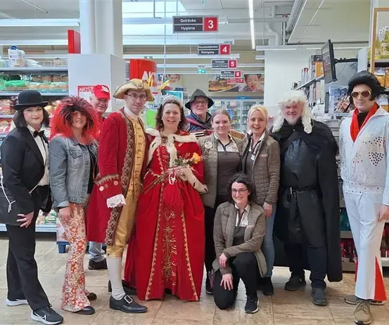 Eine Gruppe von Menschen in verschiedenen Kostümen, darunter eine Frau in einem roten Kleid, posiert für ein Foto in einem Supermarktgang.