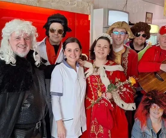 Eine Gruppe von Menschen in verschiedenen Kostümen und Perücken posiert für ein Foto in einem Raum mit roter Wand. Eine Frau in einem roten Kleid hält einen Blumenstrauß.