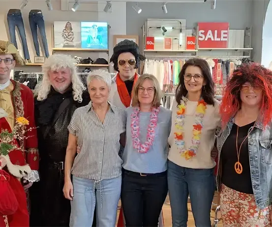 Eine Gruppe von Menschen in verschiedenen Kostümen steht in einem Bekleidungsgeschäft, lächelt und posiert für ein Foto. Hinter ihnen hängen Kleidungsstücke und Schilder mit Verkaufsinformationen.