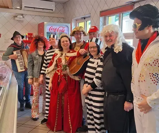 Eine Gruppe von Menschen in Kostümen steht vor einer Fleischtheke. Eine Person spielt Akkordeon, während andere lächeln und für ein Foto posieren. Coca-Cola-Automaten sind im Hintergrund.
