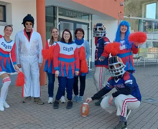 Eine Gruppe von Menschen in Cheerleader-Uniformen und einer Person in einer Football-Spieler-Uniform posiert für ein Foto vor einem Gebäude.