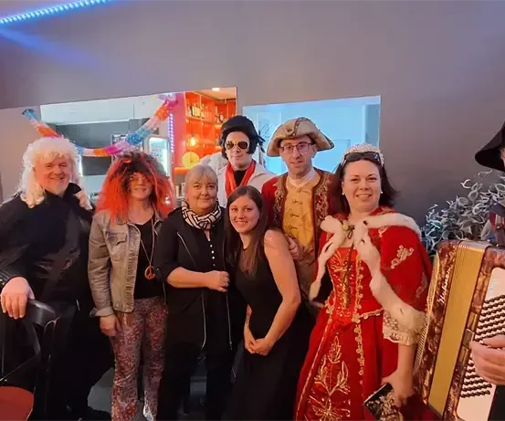 Eine Gruppe von Menschen, einige in Kostümen, posiert für ein Foto in einem Raum mit Tisch und Stühlen. Eine Person spielt Akkordeon.