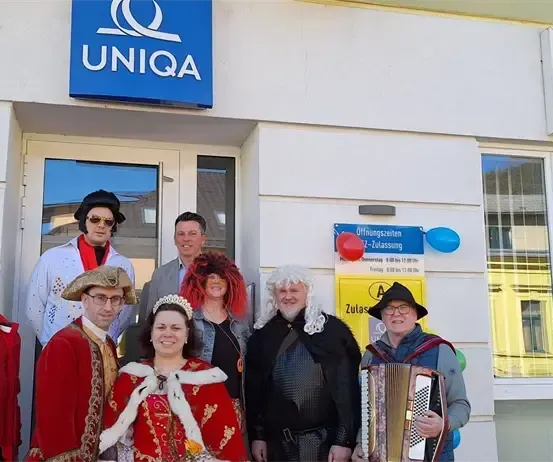 Eine Gruppe von Menschen in Kostümen posiert vor einem Gebäude mit einem UNIQA-Schild. Sie lächeln und scheinen bereit für ein Foto zu sein. Hinter ihnen sind Glastüren und Fenster.
