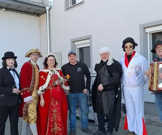 Eine Gruppe von Menschen in Kostümen, darunter eine Frau in einem roten Kleid, steht vor einem Gebäude. Ein Mann spielt Akkordeon.