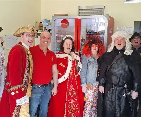 Eine Gruppe Erwachsener in Kostümen, die lächelnd und posierend für ein Foto in einem Raum mit einem Coca-Cola-Automaten im Hintergrund stehen.