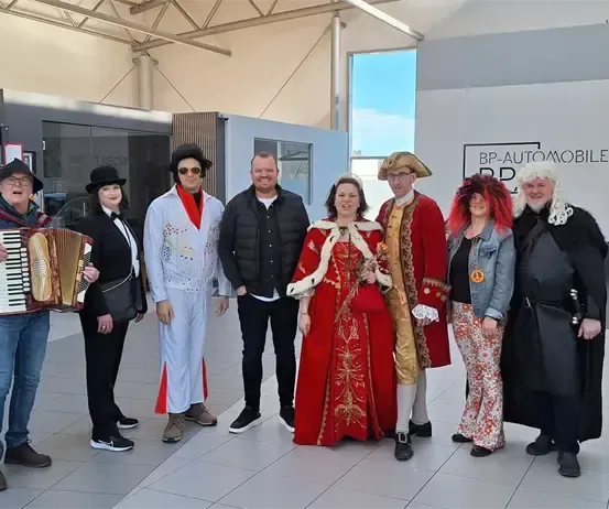 Eine Gruppe von Menschen in verschiedenen Kostümen posiert in einer Ausstellungshalle mit Autos im Hintergrund.