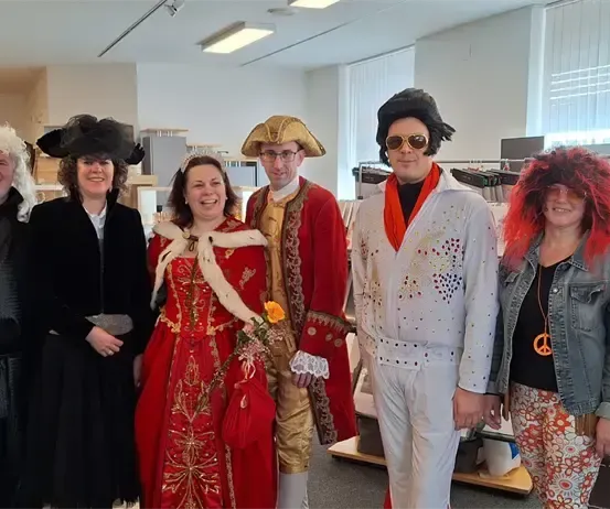 Eine Gruppe von Menschen in Kostümen, darunter ein Pirat, eine Königin, Elvis und andere, posiert für ein Foto in einem Raum.