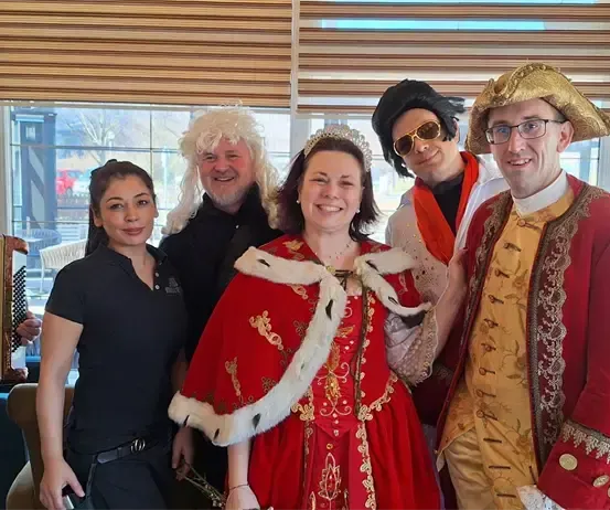 Eine Gruppe von Menschen in historischen Kostümen posiert für ein Foto, darunter eine Frau in einem roten Kleid und ein Mann, der Akkordeon spielt.