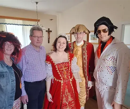 Sechs Menschen in verschiedenen Kostümen posieren für ein Foto in einem Raum mit weißen Wänden und Holzböden. Eine Frau in einem roten Kleid steht zwischen zwei Männern in Kostümen.