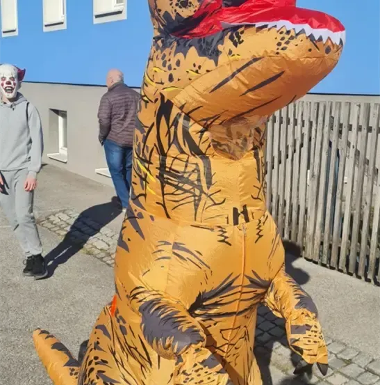 Eine Person in einem großen aufblasbaren Dinosaurierkostüm steht vor einem blauen Gebäude. Eine weitere Person mit Clownsmaske und eine dritte Person in einer Jacke sind in der Nähe.