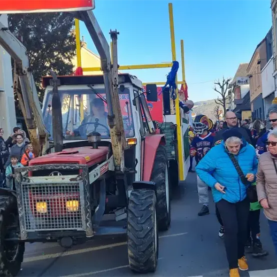 Ein Traktor führt eine Parade an, mit Zuschauern auf der Straße, darunter eine ältere Frau in einem blauen Mantel. Der Traktor hat einen roten Körper und gelbe Scheinwerfer.
