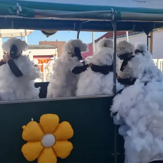 Vier Personen in flauschigen Kostümen sitzen in einem überdachten Fahrzeug mit einer gelben Blume davor. Sie posieren für ein Foto auf einer Straße.