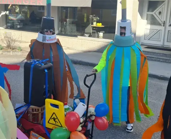 Vier Personen in Ritterkostümen stehen auf der Straße und schieben einen Wagen mit einem Gepäckstück und verschiedenen Gegenständen. Sie befinden sich vor einem Geschäft mit Glastüren und einem Schild, auf dem 'PILINGER' steht.