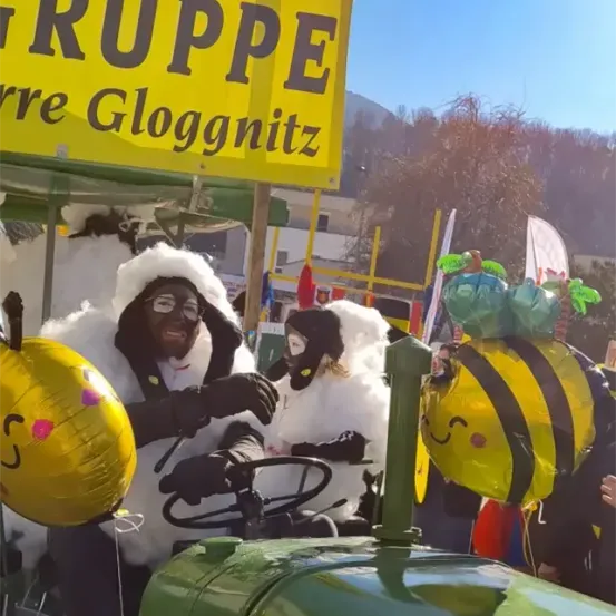 Menschen in Schafkostümen fahren während einer Parade auf einem Traktor, mit einem gelben Schild darüber und Luftballons im Vordergrund.
