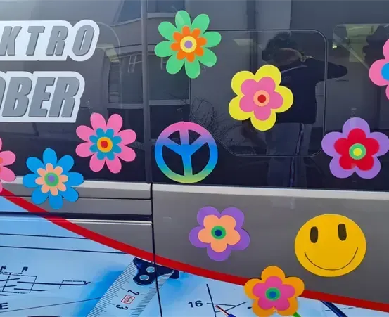 Ein Fahrzeugfenster zeigt bunte Blumen, ein Friedenszeichen und ein lächelndes Gesicht. Das Wort 'Elektro' ist sichtbar, was wahrscheinlich auf den Fahrzeugtyp hinweist.