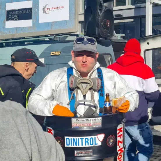 Ein Mann in Schutzkleidung trinkt ein Bier, während er in einem Fass mit der Aufschrift DINITROL steht, mit einem anderen Mann in einem roten Hut im Hintergrund.