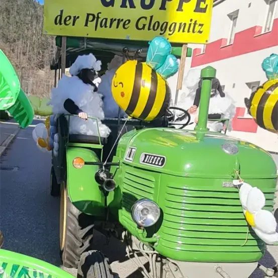 Ein Traktor, geschmückt mit Luftballons und Kostümen, fährt die Straße hinunter, mit einem Schild, das 'LEITER GRUPPE der Pfarrer Gloggnitz' lautet. Dahinter ist ein Gebäude mit rotem und weißem Rand und einem Balkon zu sehen.