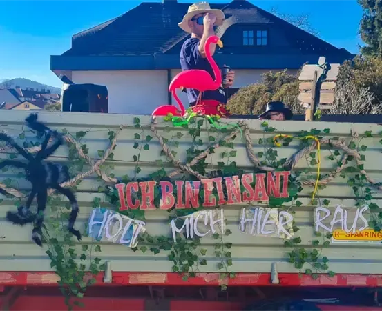 Ein dekorierter LKW mit deutschen Texten 'ICH BIN EINSAN' und 'HOLZ MICH HIER RAUS'. Ein Mann in einem Hut hält eine Flamingo-Figur. Ein Haus mit blauem Dach ist im Hintergrund.