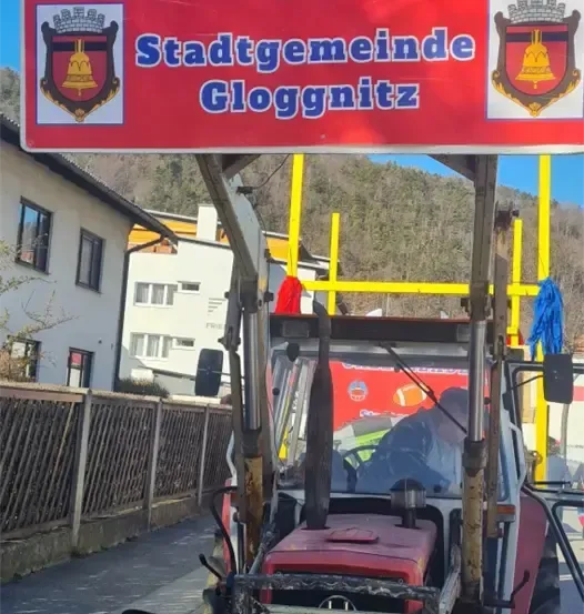 Ein roter Traktor mit einem Schild, das Cheerleader Team der Stadtgemeinde Gloggnitz steht auf der Straßenseite.