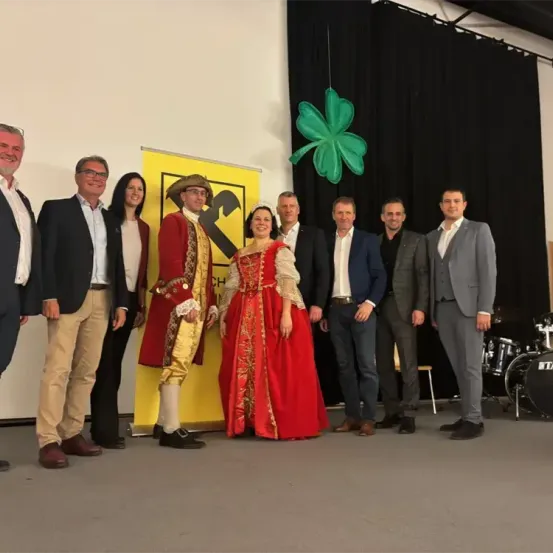 Eine Gruppe von formell gekleideten Personen steht zusammen mit einem Paar in historischen Kostümen. Sie stehen vor einem gelben Banner mit einem schwarzen Symbol. Dahinter befindet sich ein schwarzer Vorhang mit einem grünen Kleeblatt.