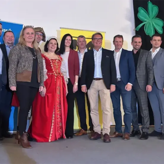 Eine Gruppe von Menschen in formeller Kleidung steht vor einer Wand mit einem grünen Kleeblatt und lächelt für ein Foto. Dahinter ist ein blauer Banner mit weißem Text zu sehen.