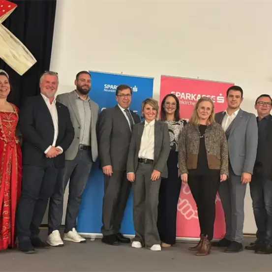Eine Gruppe von Menschen in verschiedenen Kleidungsstücken steht vor einer weißen Wand, einige in formeller Kleidung. Ein Banner mit dem Wort 'Sparkasse' befindet sich hinter ihnen. Eine pilzartige Dekoration ist auf der linken Seite.