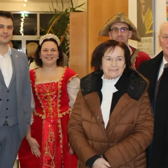 Eine Gruppe von Menschen, darunter eine Frau in einem roten Kleid, ein Mann in einem Anzug und eine ältere Frau in einer braunen Jacke, posieren für ein Foto.