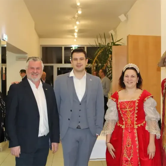 Fünf Personen posieren für ein Foto in einem Raum mit einer Holzwand, einer Pflanze und Lampen. Sie tragen formelle und traditionelle Kleidung.