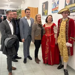 Fünf Personen stehen eng beieinander in einem Flur, zwei von ihnen sind in Kostümen gekleidet. Sie scheinen zu lächeln und für ein Foto zu posieren. Hinter ihnen hängen Poster an der Wand.