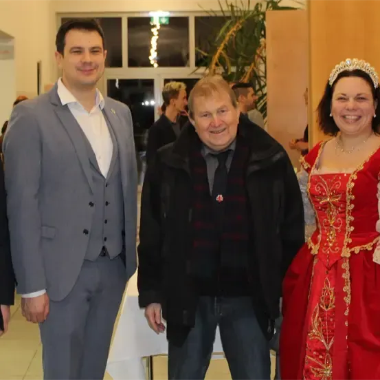 Fünf Menschen stehen in einem Raum, lächeln und posieren für ein Foto. Der Mann auf der linken Seite trägt einen Anzug, und die Frau auf der rechten Seite trägt ein rotes Kleid. Der Mann in der Mitte trägt eine schwarze Jacke und eine Halskette. Im Hintergrund sind weitere Menschen zu sehen.